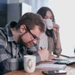 A couple sits at a kitchen table, reviewing finances with a laptop and calculator.